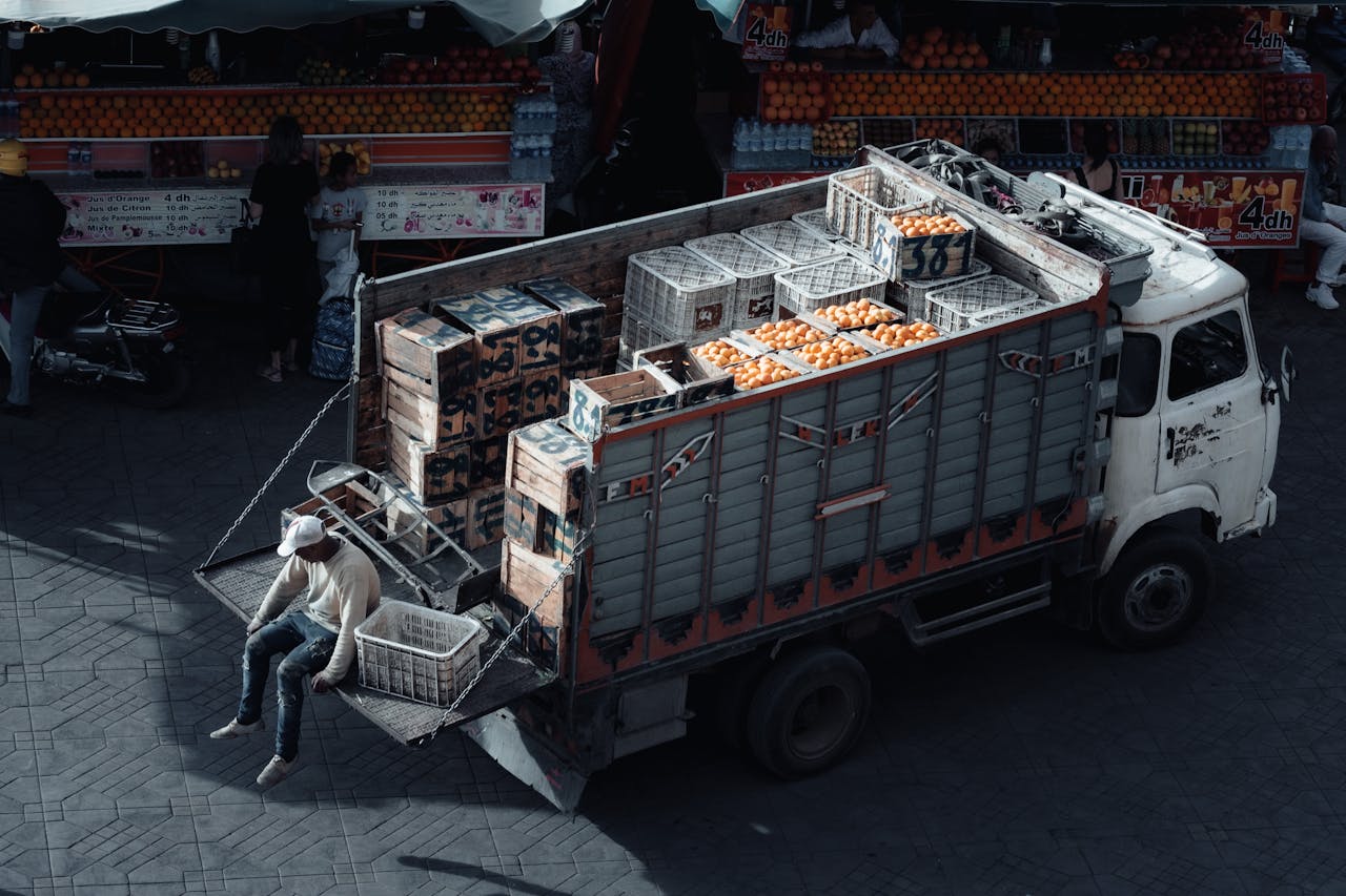 services-01 A truck loaded with crates of oranges parked at an urban market scene during daytime.